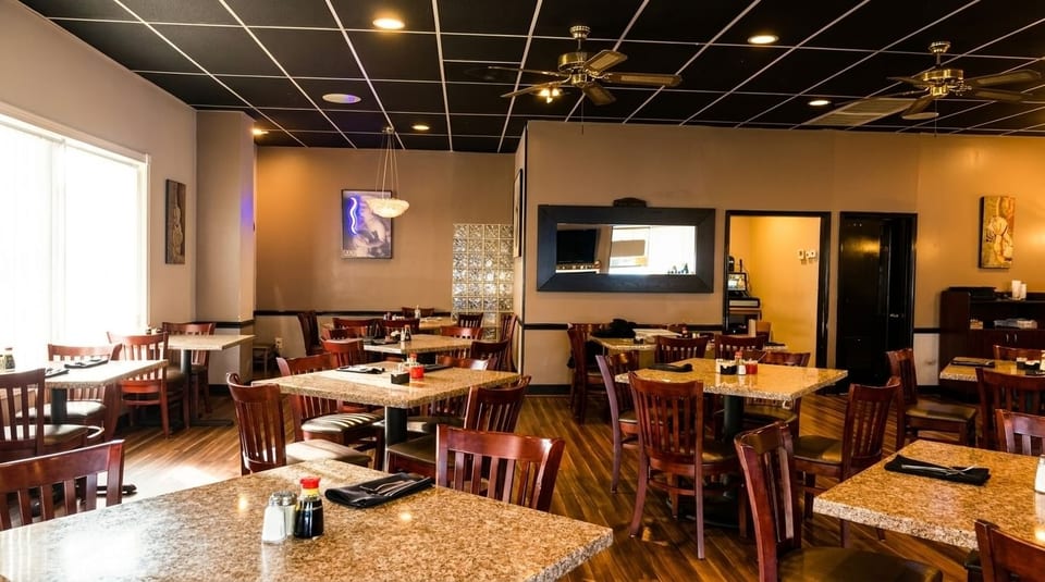 Empty restaurant dining room with granite-topped tables, dark wooden chairs, and a black ceiling with ceiling fans.